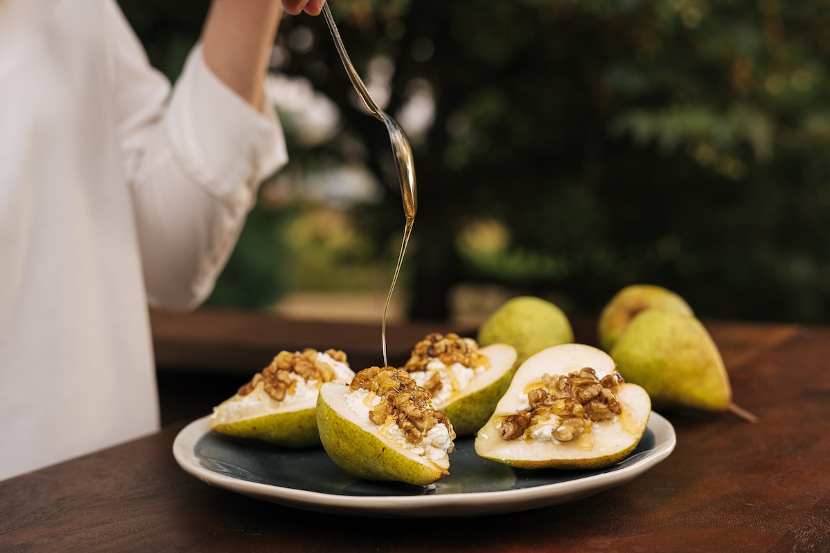 Four Warmed Pear Halves on a Plate Stuffed with Chèvre and Nuts with Chef Drizzling Honey Over Top with a Spoon