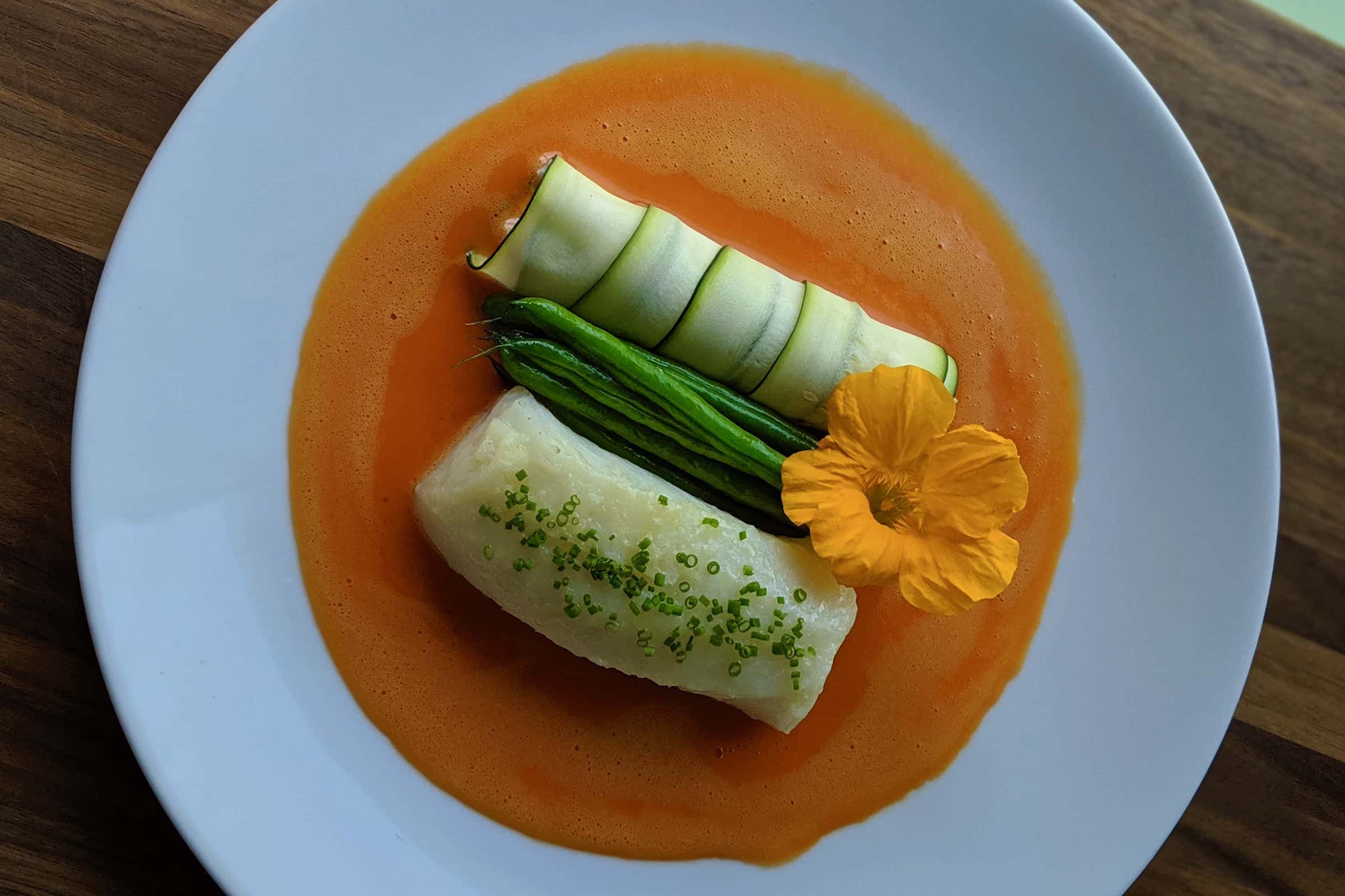 Elegant plate with cod filet, green beans and paper thing zucchini sheets wrapping ricotta cheese in a red pepper sabayon sauce